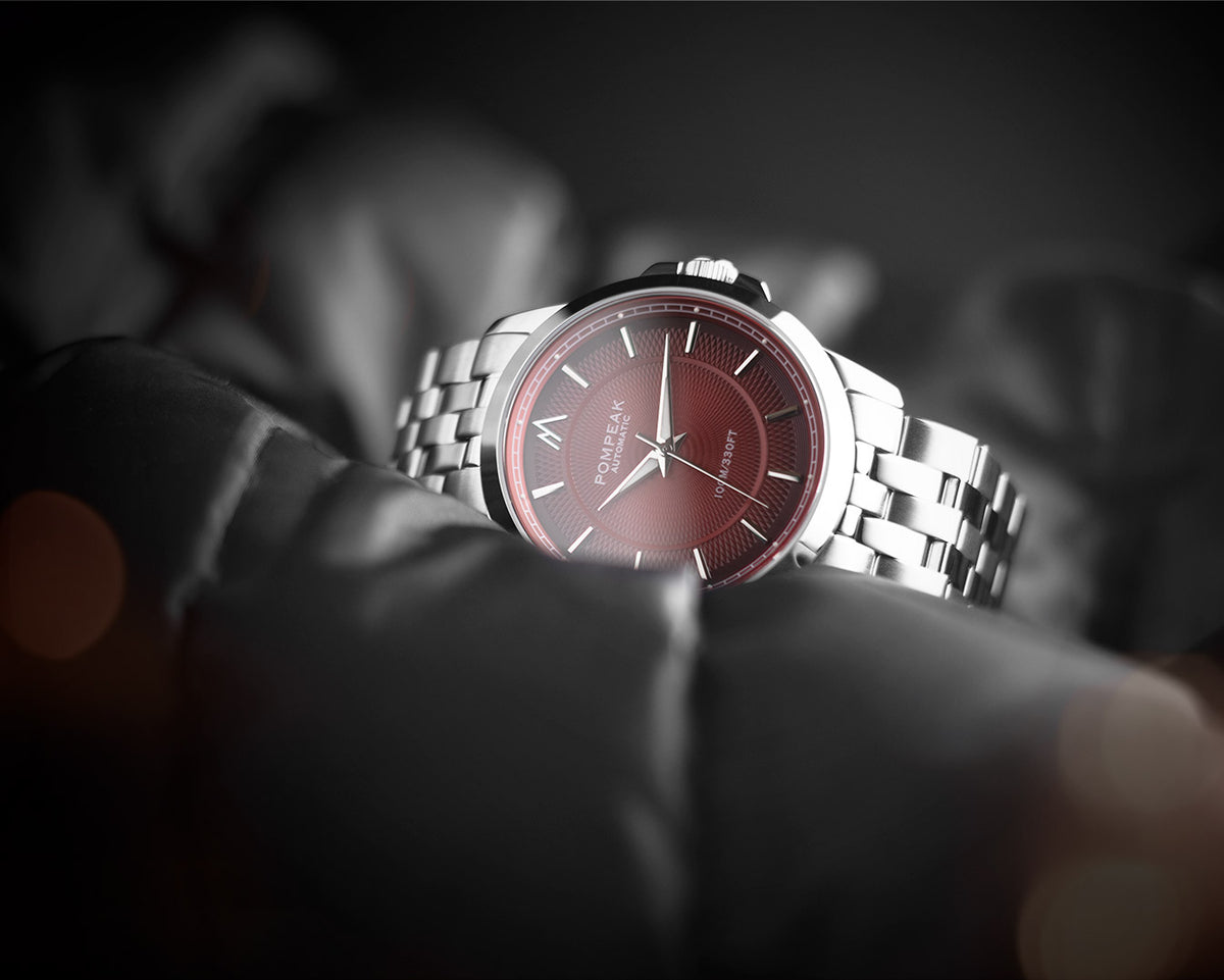 A close-up of a wristwatch with a silver metal bracelet and a dark red dial, resting on a dark, blurred surface. The brand name Pompeak is visible on the dial.