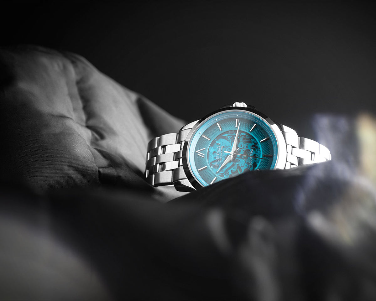 A silver metal bracelet watch with a vibrant turquoise blue dial, resting on a dark, textured fabric surface against a dark background.