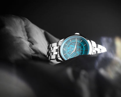 A silver metal bracelet watch with a vibrant turquoise blue dial, resting on a dark, textured fabric surface against a dark background.